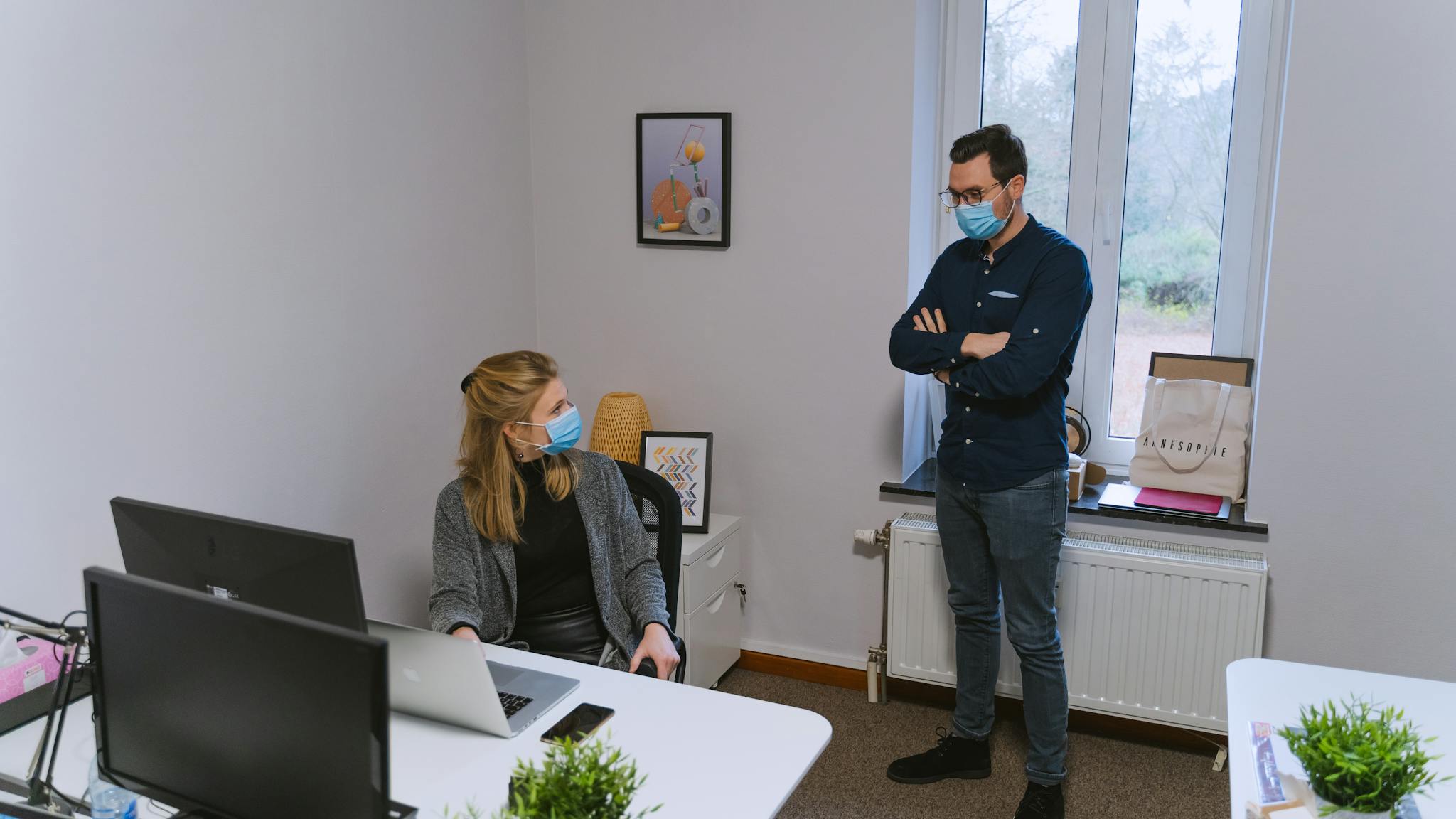 Two colleagues discussing in an office while wearing face masks, emphasizing pandemic safety.