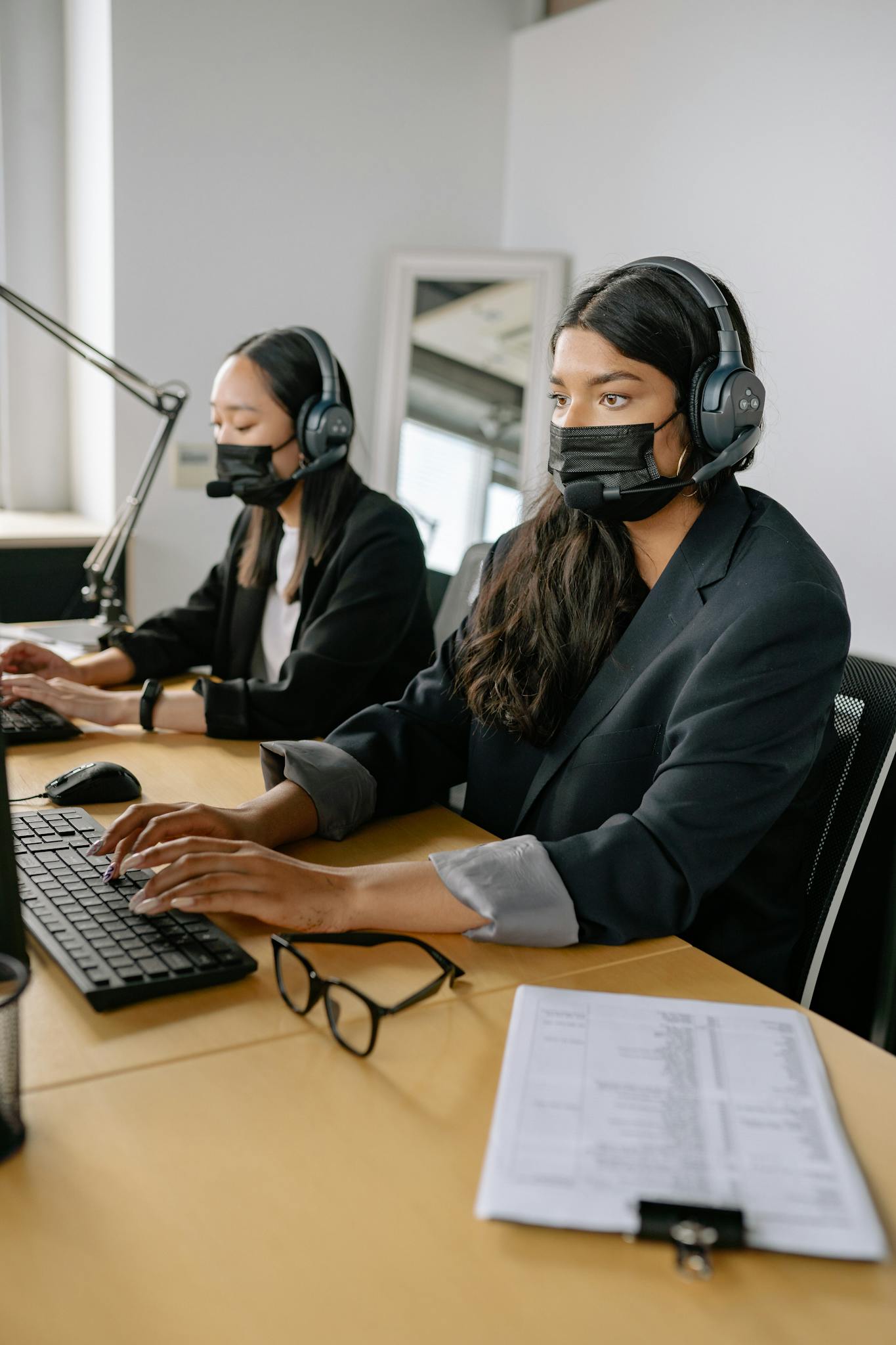 Two women in headsets and masks working at a call center office desk.